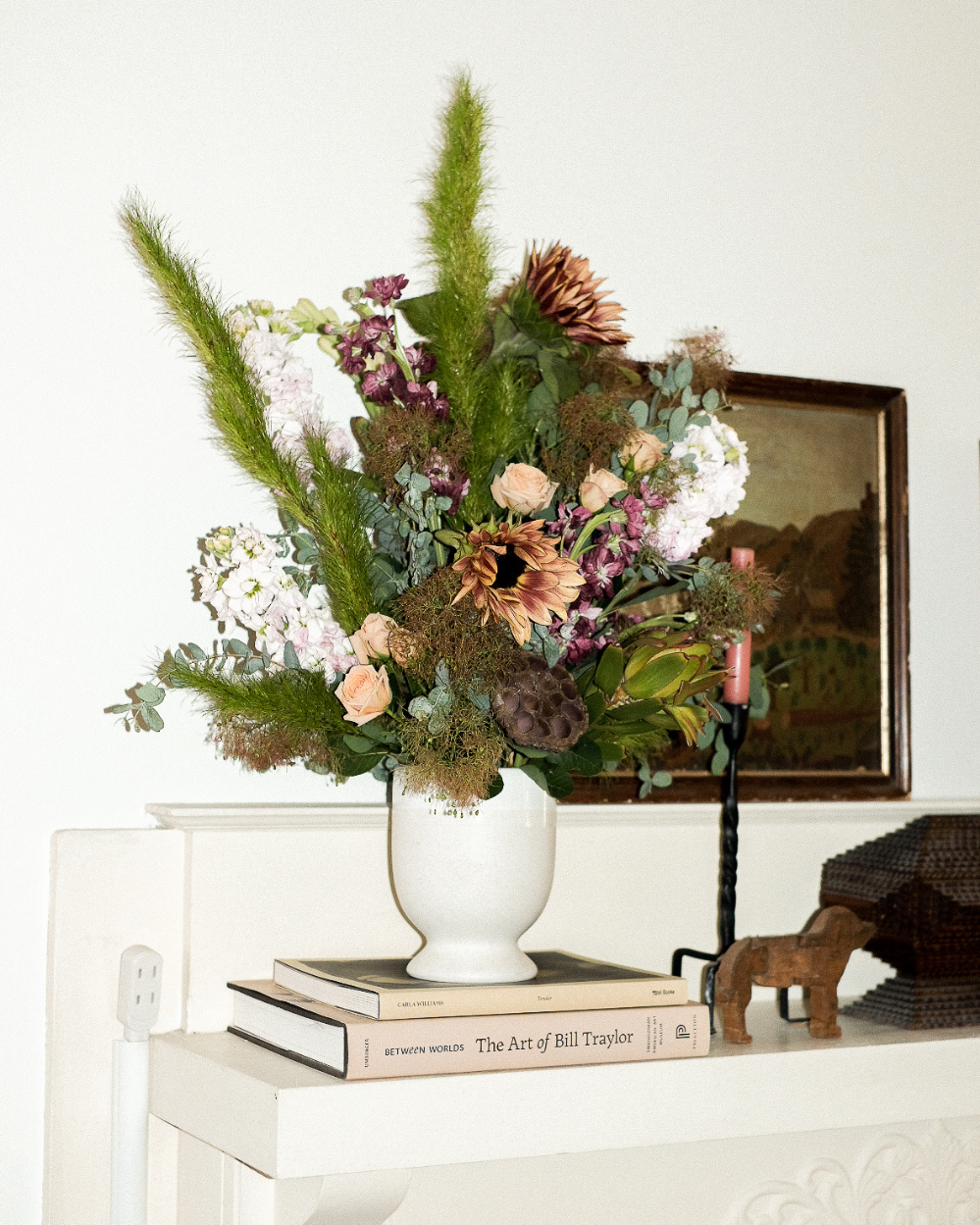 A dried and floral arrangement in a white ceramic vase, styled on a white mantelpiece alongside books, a small wooden elephant figurine, and a framed picture against a white wall.