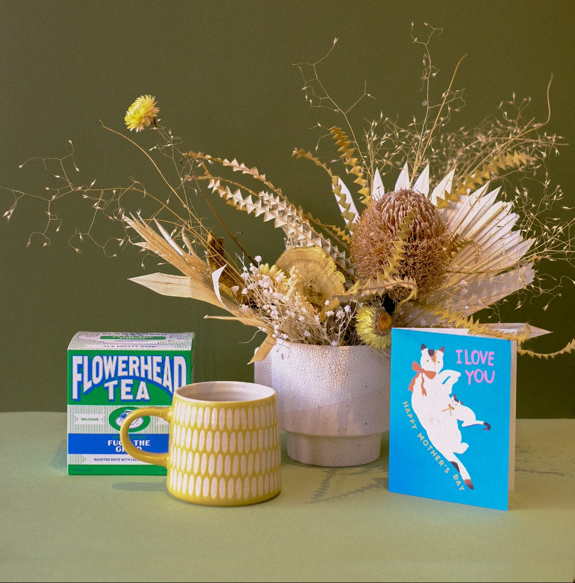 Decorative arrangement with a potted plant, tea box, and greeting card on a green surface.