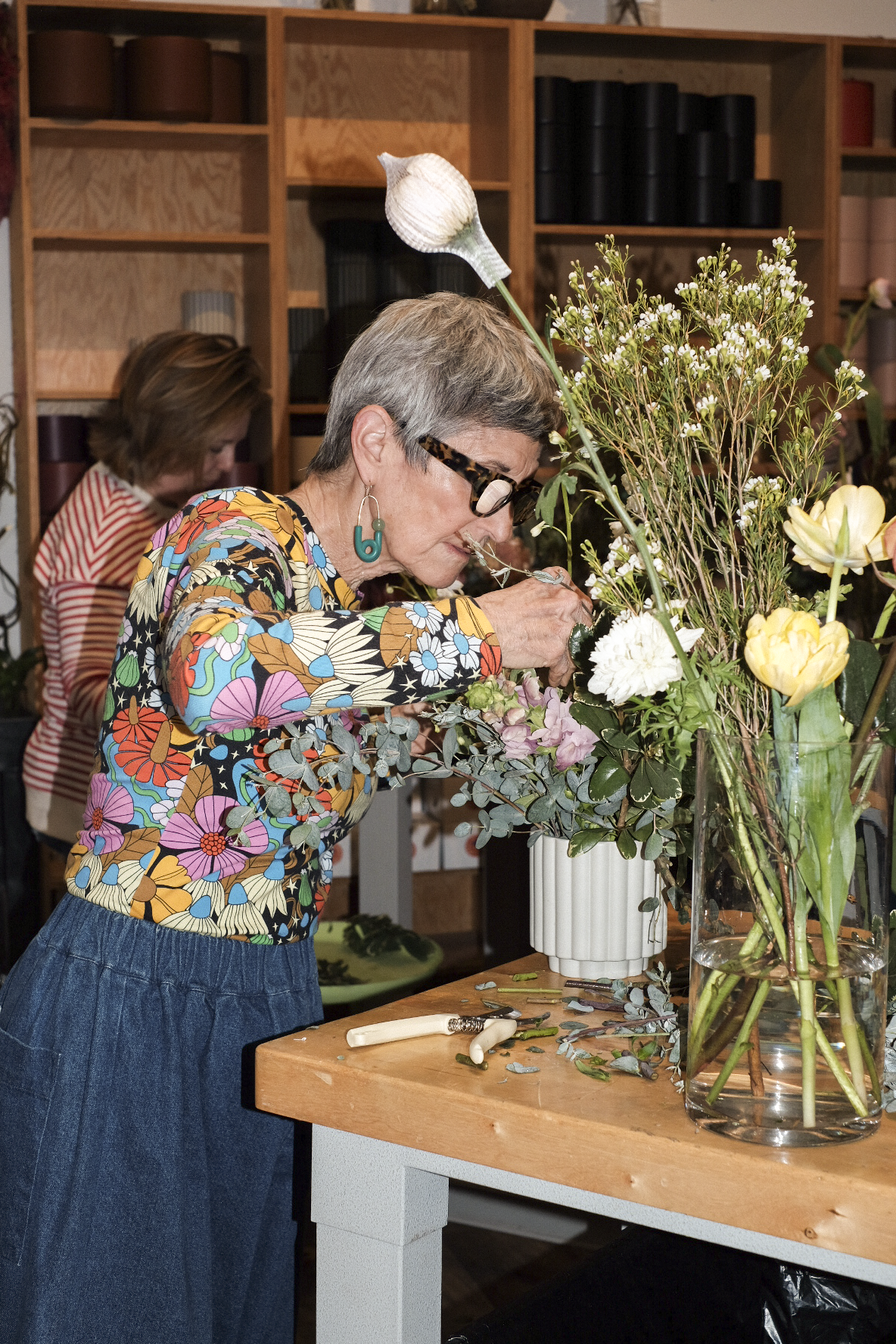 Woman arranging flowers in a vase at a wooden table with a bookshelf in the background.