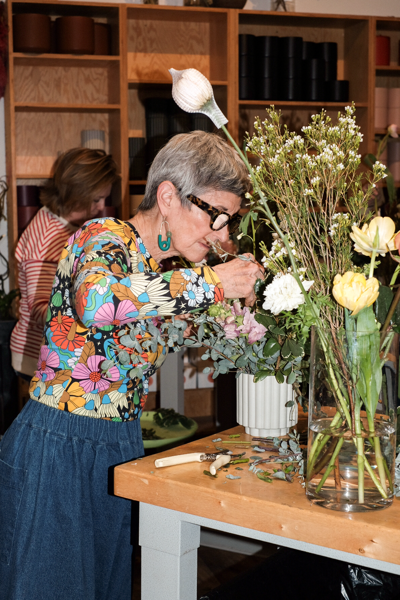 Woman arranging flowers in a vase at a wooden table with a bookshelf in the background.